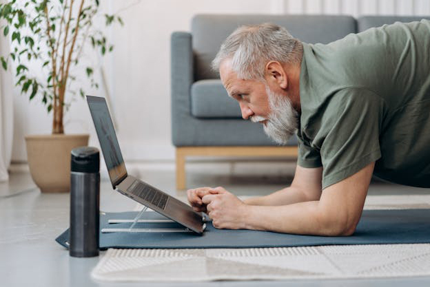older man looka at laptop while doing yoaga pose on mat